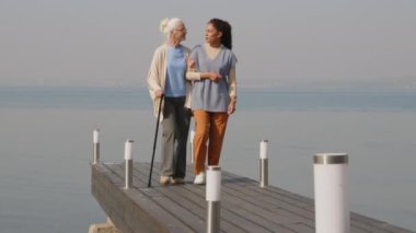 Genç bir kadın bakıcı, yaşlı bir kadınla sabah birlikte göl kıyısında yürürken konuşuyor.