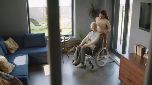 リビングルームを通して車椅子を押し 自宅で彼女を助けながらシニア女性と話す女性介護者のハイアングルショット — ストック動画