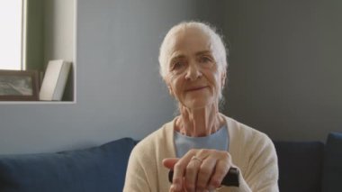 Yaşlı kadın portresi, gri saçlı, baston üzerinde el ele tutuşan ve evdeki kanepede otururken kameraya poz veren.
