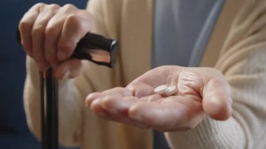 Bir elinde baston, diğerinde hap tutan yaşlı bir kadın resmi.