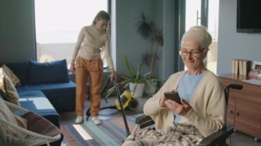 Yaşlı kadın tekerlekli sandalyede oturuyor ve akıllı telefon kullanıyor. Hemşireye oturma odasını süpürmesi ve evi temizlemesi için yardım ediyor.