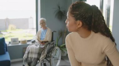 Tekerlekli sandalyedeki yaşlı kadın arka planda kitap okurken Afrikalı Amerikalı kadın hemşire yardımcısının yerleri süpürdüğü ve evi toparladığı sahneyi çek.