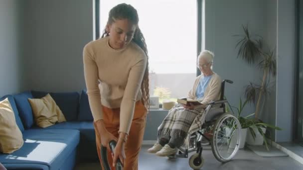 リビングルームの床にカーペットを掃除する若い女性の介護者のショットを傾けて 車椅子の年配の女性を家のクリーニングで助けながら — ストック動画