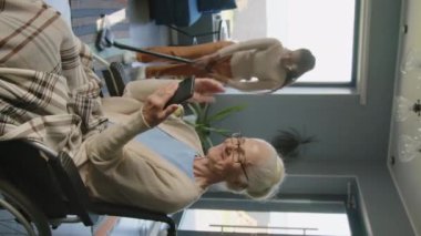 Tekerlekli sandalyede oturan yaşlı bir kadın ve akıllı telefondan arama yaparken oturma odasını süpüren bir hemşirenin dikey görüntüsü.