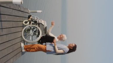 Tekerlekli sandalyedeki kadın bakıcı ve yaşlı kadının şafak vakti tahta iskelede dururken manzaralı göl manzarasını tartışırken dikey çekimi.