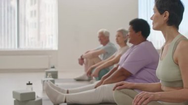 Bir grup yaşlı kadın ve erkeğin spor salonunda oturup yoga hocasıyla pratik yaparken sohbet ettiği fotoğraflara odaklan.