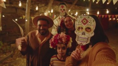 Geceleri arka bahçede Dia de Muertos kutlamaları sırasında el yapımı kağıt kafatası maskelerinin altında yüzlerini saklayan ve kameraya gülümseyen mutlu Meksikalı aile portresi.