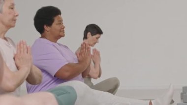 Spor kıyafetleri içinde bir grup olgun kadın yoga stüdyosunda gözleri kapalı ve elleri Namaste 'de meditasyon yaparken oturuyorlar.