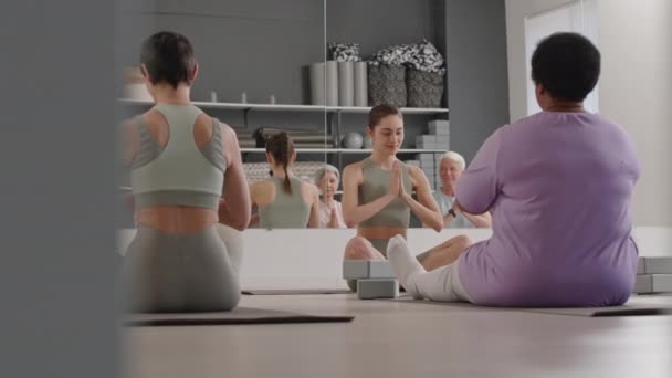 若い女性ヨガインストラクターとスタジオで床に座っているスポーツウェアのシニアの人々のグループ 瞑想を実践しながら祈りの位置と目を閉じる — ストック動画