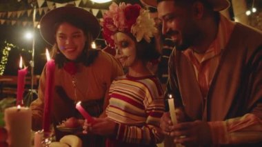 Yarı boyalı iskeleti olan Meksikalı küçük bir kızın arka bahçede ailesiyle Dia de Muertos 'ta sunak başında mum yakarken orta boy bir fotoğrafı.