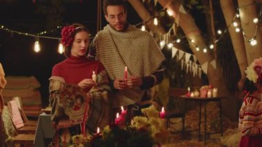 Geleneksel kıyafetleri olan, elinde mumlarla sunak başında duran ve arka bahçede Ölüler Günü kutlamaları sırasında sohbet eden Meksikalı genç çiftin orta boy fotoğrafı.