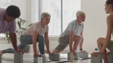 Stüdyoda kadın eğitmenle antrenman yaparken yoga bloklarıyla hamle yapan bir grup yaşlı insan.