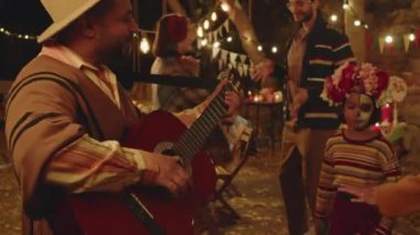 Neşeli Meksikalı bir adamın gitar çaldığı orta boy bir fotoğraf ve diğer insanlar geceleri arka bahçede dans ederken Dia de Muertos ailesinin kutlamaları sırasında.