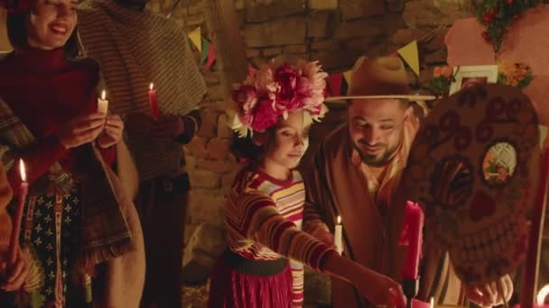 伝統的なお祝いの衣装で陽気な大きなヒスパニック系家族のショットをズームアウト キャンドルで儀式を行いながら 死者の日にクラフトスケルトンや頭蓋骨で装飾された祭壇で立っています — ストック動画
