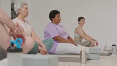 Spor giyimli bir grup olgun kadın yoga stüdyosunda egzersiz minderlerinin üzerinde oturuyor ve antrenmandan önce eğitmenle konuşuyorlar.
