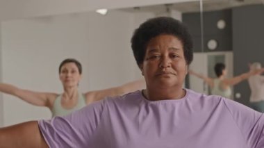 Namaste 'de el ele tutuşan ve yoga stüdyosunda grup meditasyonu yaparken kameraya poz veren olgun Afro-Amerikan kadın portresi.