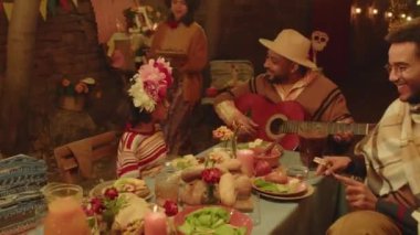 Arka bahçede Dia de Muertos 'ta neşeli, İspanyol bir ailenin açık havada akşam yemeği yediği orta boy bir fotoğraf. Yemek servisi yapan kadınlar ve gitar çalan erkekler.
