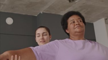 Genç bayan yoga öğretmeninin fotoğrafına yakınlaşın. Kıdemli Afrikalı Amerikalı kadınla gülümseyip konuşurken stüdyoda savaşçı pozu verirken ona yardım edin.