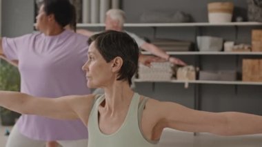 Kıdemli kısa saçlı, spor giyimli kadın grup yoga dersi sırasında savaşçı ve üçgen pozları veriyor.