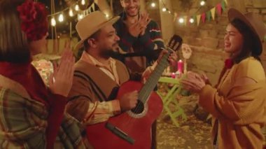 Meksikalıların, Ölüler Günü 'nde aile üyeleri ve arkadaşlarıyla gitar çalarken arka bahçede mumlar ve kadife çiçekleriyle süslenmiş dans edip şarkı söylerken çekilmiş orta boy bir fotoğraf.
