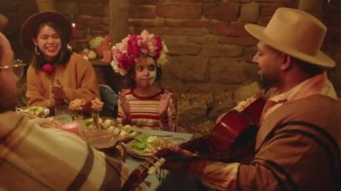 Deve şapkası ve panço takmış İspanyol bir adamın ailelerine gitar çalması ve gece rahat arka bahçede yemek masasında oturup Ölülerin Günü 'nü kutlaması.