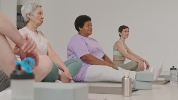 ヨガスタジオでエクササイズマットに座って練習前にインストラクターと話すスポーツウェアの成熟した女性のグループ — ストック動画
