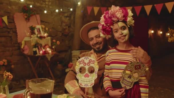 ヒスパニック系男性と彼の小さな娘の半分塗られた顔と花輪は 死者の日に裏庭に手作りの紙の頭蓋骨マスクでカメラのためにポーズします — ストック動画