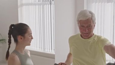 Spor salonundaki genç bayan fitness eğitmeninin yardımıyla dambıl yumruklayan neşeli, gri saçlı yaşlı adamın orta boy fotoğrafı.