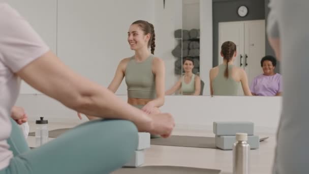 フィットネススタジオで足で座っている若い美しい女性ヨガの先生は 練習前に女性のグループと笑顔と話す — ストック動画