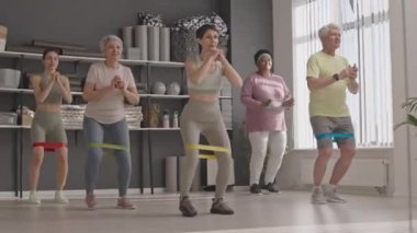 Bir grup yaşlı insan, kadın fitness koçunun kontrolü altında spor yaparken aynanın karşısına geçip direniş bandı adımları atıyor.