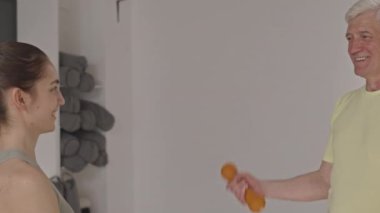 Genç bayan kişisel antrenör ve pozitif kıdemli adamın spor yaparken halterli kıvrımlar yapıp sohbet etmelerinin arka plan çekimi.