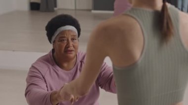 Kıdemli Afrikalı Amerikalı bir kadının egzersiz topunun üzerinde el ele tutuşup spor salonunda spor yaparken spor eğitmeniyle konuşması.