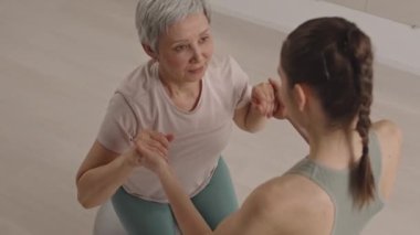 Yüksek açılı, yaşlı, gri saçlı bir kadının egzersiz topuna oturduğu ve spor salonunda antrenman yaparken kadın fitness eğitmeninin yardımıyla ayağa kalktığı bir fotoğraf.