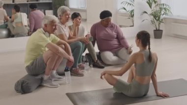 Antrenmandan sonra dinlenen, su içen ve önlerinde oturan kadın fitness hocasıyla sohbet eden bir grup yaşlı insan.