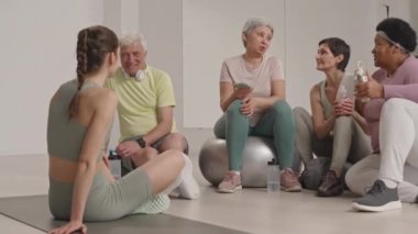 Bir grup pozitif yaşlı arkadaş spor yaptıktan sonra dinleniyorlar ve spor salonunda önlerinde oturan bayan fitness hocasıyla sohbet ediyorlar.