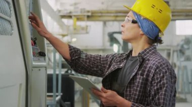 Baret ve güvenlik gözlüklü kadın işçinin taş kesme fabrikasındaki dijital tabletle endüstriyel makineyi çalıştırmasını çek.