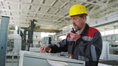 Fabrikadaki endüstriyel makineyi çalıştırırken telsiz kullanarak iş kıyafetlerinde deneyimli erkek mühendisin orta boy görüntüsü.
