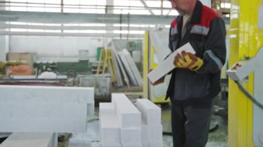Üniformalı ve eldivenli olgun bir erkek işçinin beyaz küboit mermer bloğu taşırken ve taş fabrikasında çalışırken onu üst üste koyarken orta uzunlukta bir pozu.