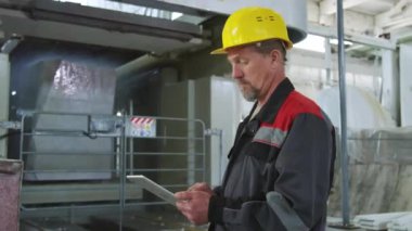 Kıdemli teknisyenlerin kasketli ve iş kıyafetli orta boy görüntüleri endüstriyel makineleri kontrol ediyor mermer blokları yıkıyor ve iş günü taş fabrikasında dijital tablet kullanıyor.