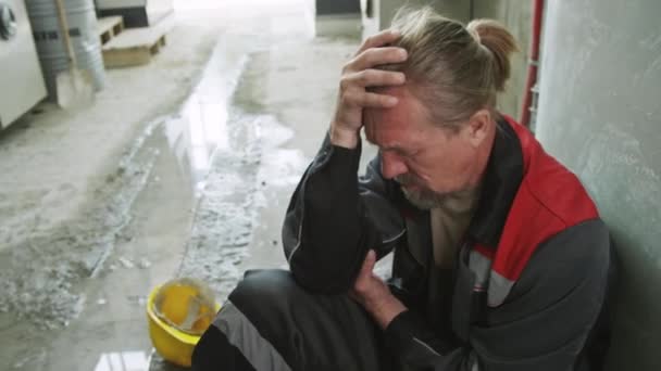 ハイアングルハンドヘルドショット 工業工場の床に座っている作業員の疲れ果てた上級男性 額にヤシを抱え ハードワークの後の頭痛に苦しむ — ストック動画