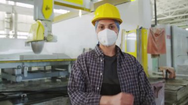 Solunum maskeli ve koruyucu kasklı kadın işçinin orta boy portresi. Kollarını kavuşturup endüstriyel fabrikada kendine güvenen kameraya poz veriyor.