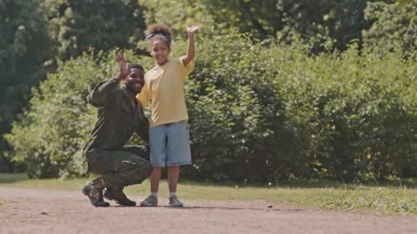 快乐的非裔美国军人身着迷彩服 他可爱的小女儿看着镜头 在阳光灿烂的日子里 一边微笑着一边挥手 一边在公园里摆姿势 — 图库视频影像