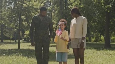 Küçük Afrikalı Amerikalı kız elinde Amerikan bayrağı, askeri üniformalı babasıyla yürüyor ve güneşli bir yaz gününde parkta genç bir anne.