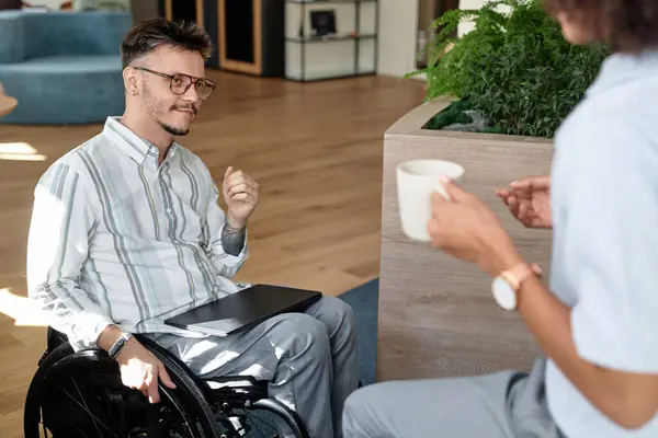 Young businessman with disability sitting in wheelchair and talking to his colleague during coffee break in office hall