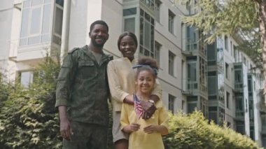 Askeri üniformalı mutlu siyah asker, karısı ve küçük kızı sokakta Amerikan bayrağı ile birlikte gülümsüyor ve poz veriyorlar.