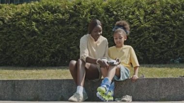 Afrika kökenli Amerikalı bir anne, kızına parkta paten kaymadan önce koruyucu dizlik takmasına yardım ediyor.