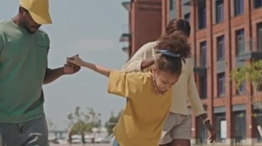 Afrikalı Amerikalı bir kızın ebeveynlerinin yardımıyla kaykay sürmeyi öğrenirken aşağı eğilin.