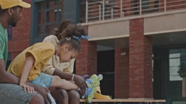 Afrika kökenli Amerikalı anne ve baba, sokakta otururken küçük kızın paten giymesine yardım ediyor.