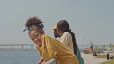Küçük Afrikalı Amerikalı bir kızın portresi nehir kenarındaki korkuluklara yaslanıyor ve güneşli bir yaz gününde ebeveynler arkada dururken kameraya gülümsüyor.