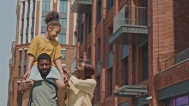 Babalarının omuzlarında gezen neşeli Afrikalı Amerikalı kızın orta boy bir fotoğrafı. Ailesiyle sokakta yürürken ve yaz günü dışarıda ailecek vakit geçirirken.